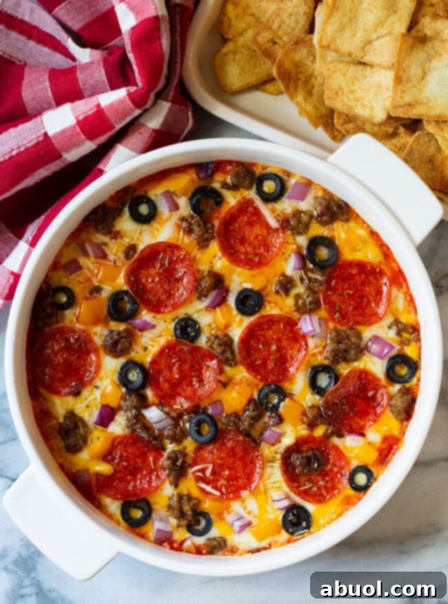 A top-down view of a white round dish, generously laden with a perfectly baked Supreme Pizza Dip. The dip is topped with glistening pepperoni slices, crumbled cooked sausage, vibrant black olive slices, and finely chopped red onion, ready for serving.