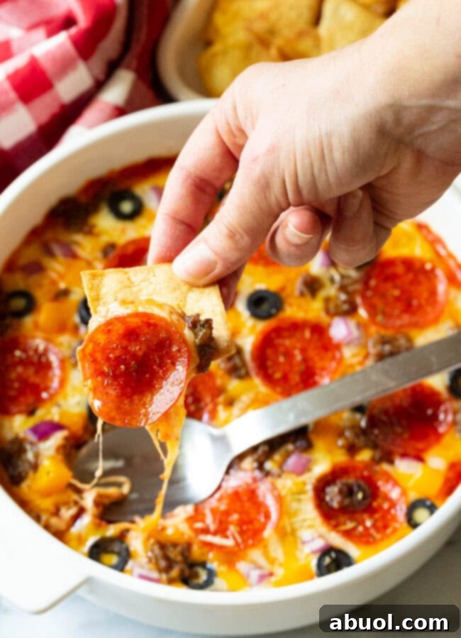 A hand skillfully uses a crisp pita chip to scoop a rich bite of pepperoni pizza dip, showcasing the melted cheese and visible toppings.
