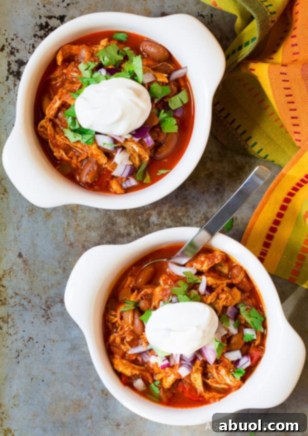 Roasted Red Pepper Crockpot Chicken Chili 4 Top down view two white bowls of shredded red pepper chicken chili topped with diced red onions, cilantro, and dollop of sour cream.