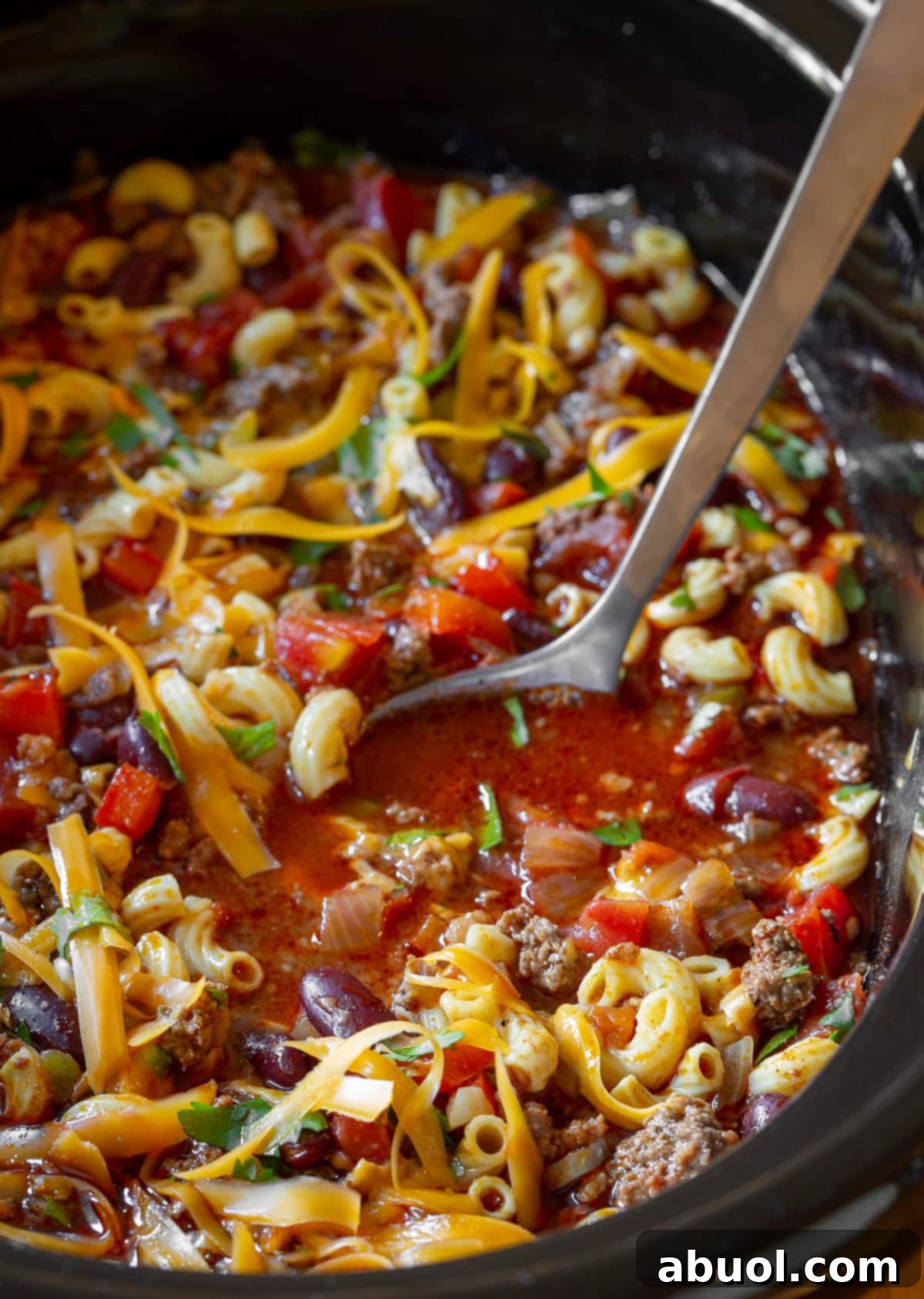 A large metal ladle serving generous portions of rich, cheesy slow cooker chili mac and cheese from a crockpot, highlighting the comforting texture and abundant ingredients.