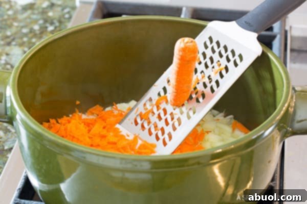 A close-up shot of a hand shredding carrots directly into a green cooking pot, where onions and garlic are already sautéing.