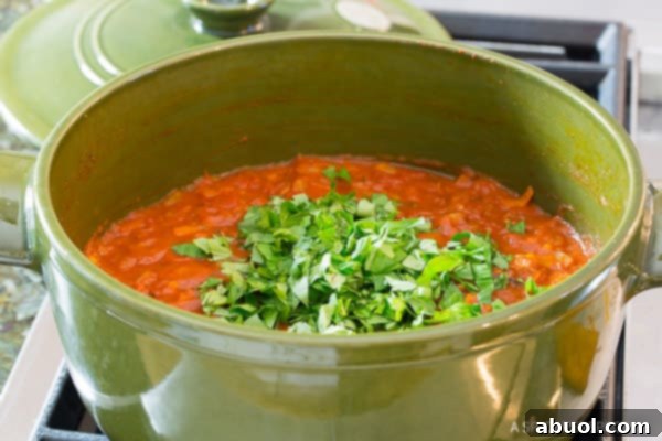 A hand gently adding fresh chopped basil to a vibrant red tomato sauce simmering in a green cooking pot, emphasizing freshness.