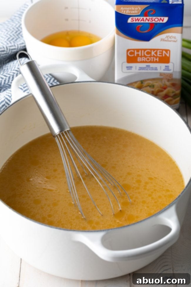 A large white pot filled with simmering chicken broth, with a metal whisk resting inside. In the background, a carton of chicken broth and a bowl of whisked eggs sit on a kitchen counter.