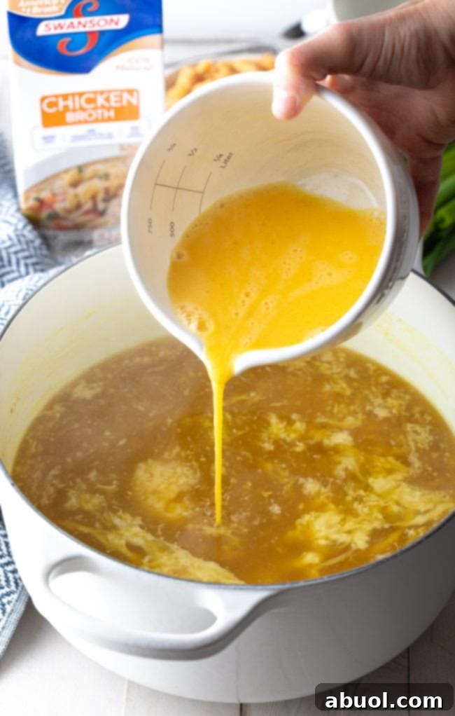 A stream of whisked eggs being slowly poured from a white measuring bowl into a pot of simmering golden broth, creating delicate egg ribbons.
