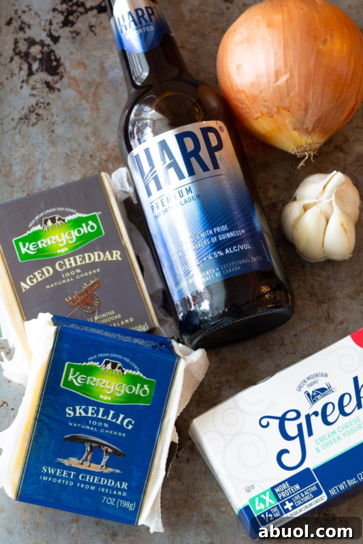 A vibrant display of fresh ingredients including a large onion, garlic cloves, butter, Harp lager, chicken broth, various Irish cheeses, cream cheese, cornstarch, and Dijon mustard, laid out on a wooden surface for making Irish Beer Cheese Soup.