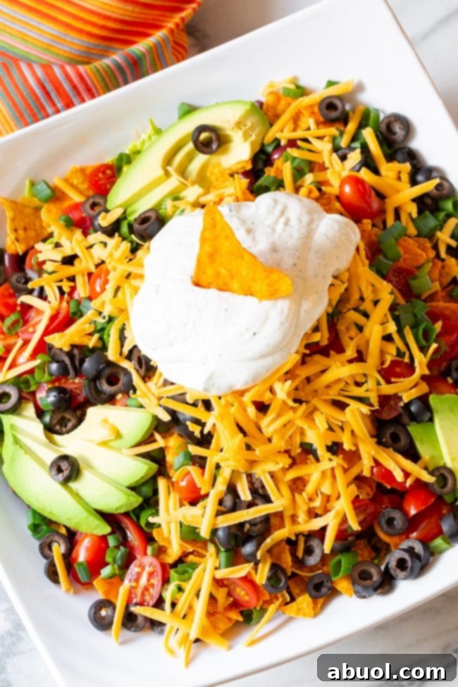 A vibrant Doritos taco salad generously loaded with fresh lettuce, halved grape tomatoes, sliced black olives, chopped green onions, creamy avocado slices, and shredded cheddar cheese. A dollop of ranch sour cream, adorned with a single Dorito chip, sits proudly in the center.