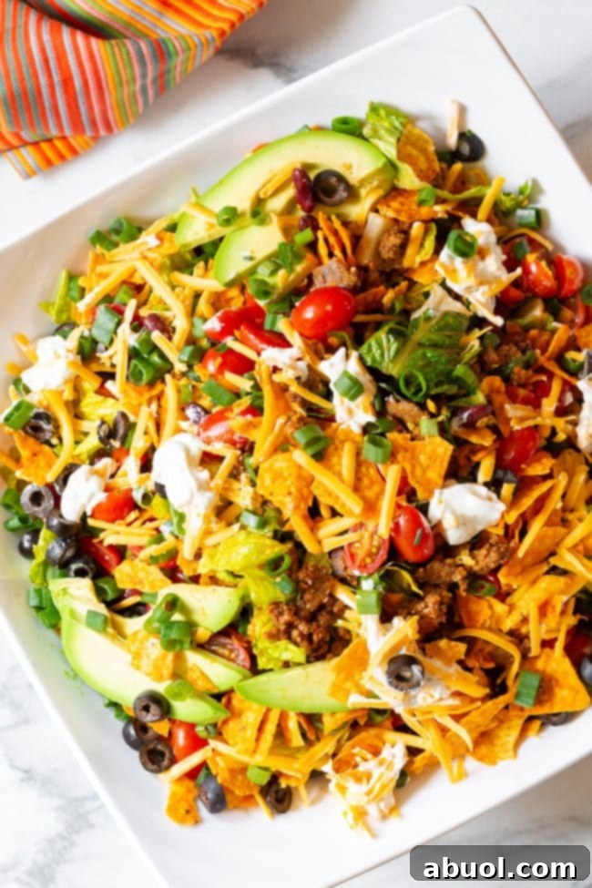 A beautifully presented Doritos taco salad, fully loaded with fresh lettuce, halved grape tomatoes, sliced black olives, chopped green onions, creamy avocado slices, and shredded cheddar cheese. Several dollops of ranch sour cream are artfully arranged around the edge.