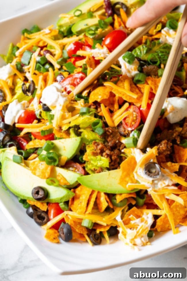 A hand using wooden tongs to toss the colorful Dorito taco salad, showing the crushed Dorito chips mixing with fresh lettuce and other salad fixings.