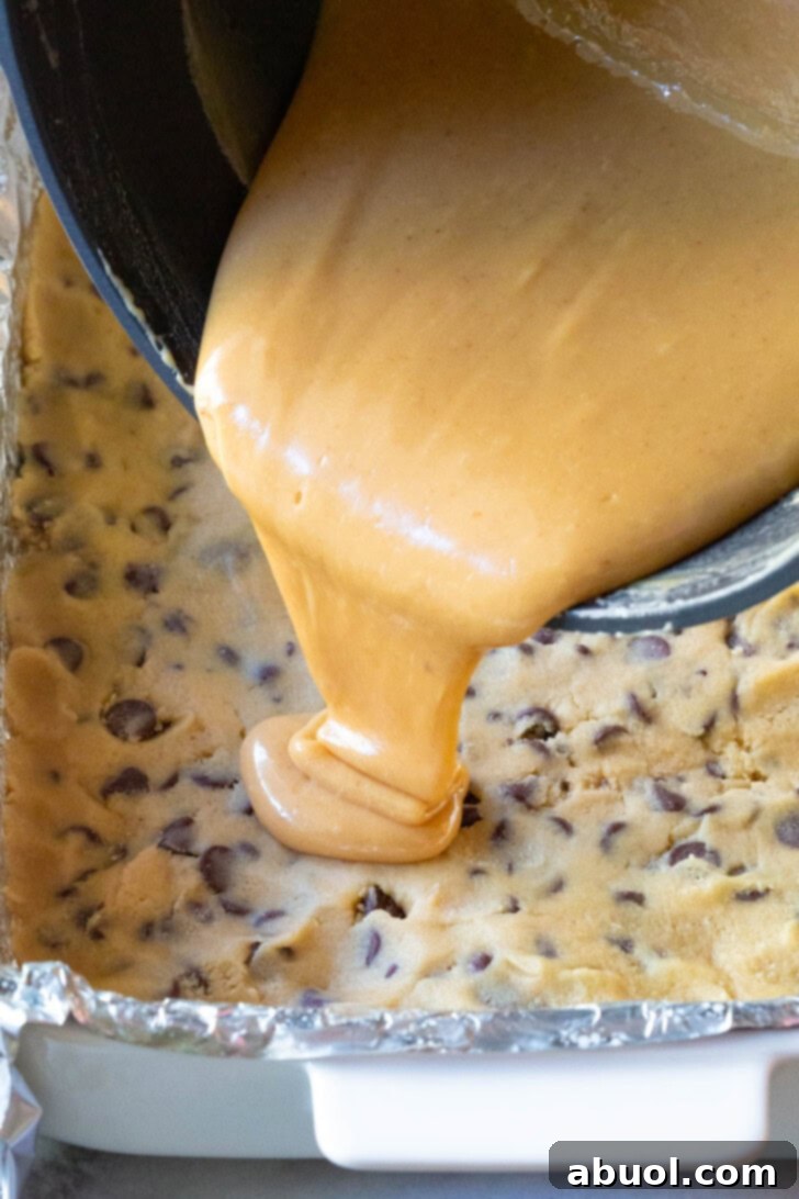 Warm caramel sauce being poured onto a layer of cookie dough in a baking dish.