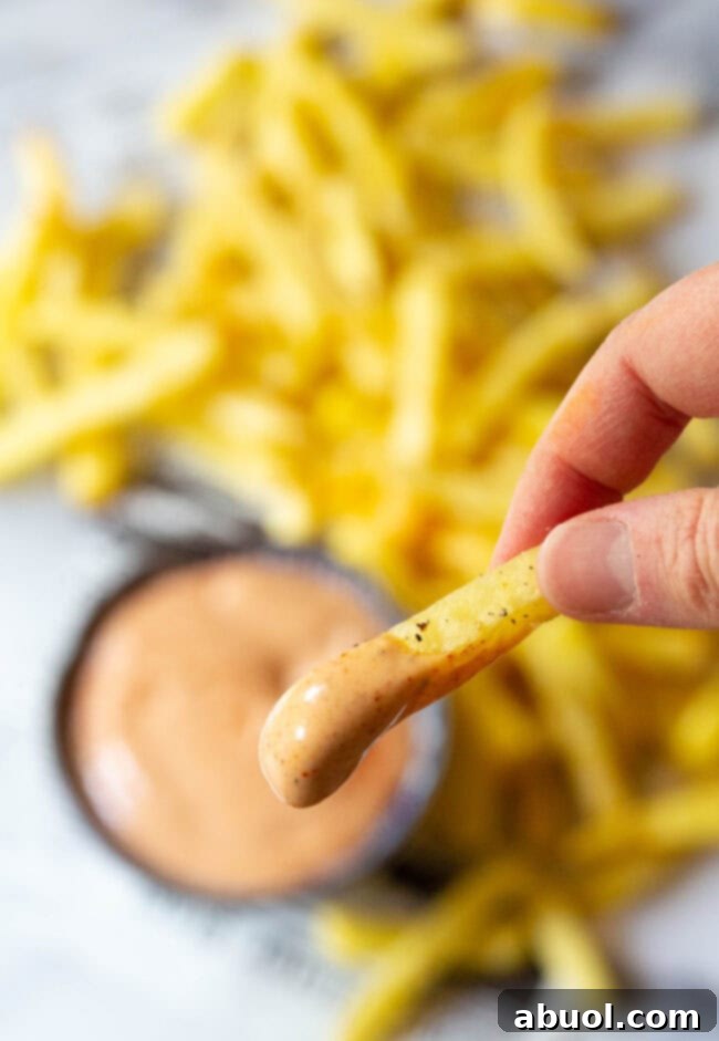 Hand holding a fry dipped in sauce with more sauce and fries in the background. 