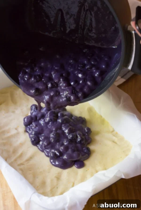 Warm, thick blueberry sauce being carefully poured from a pot onto the prepared cookie base in a baking dish, spreading evenly.