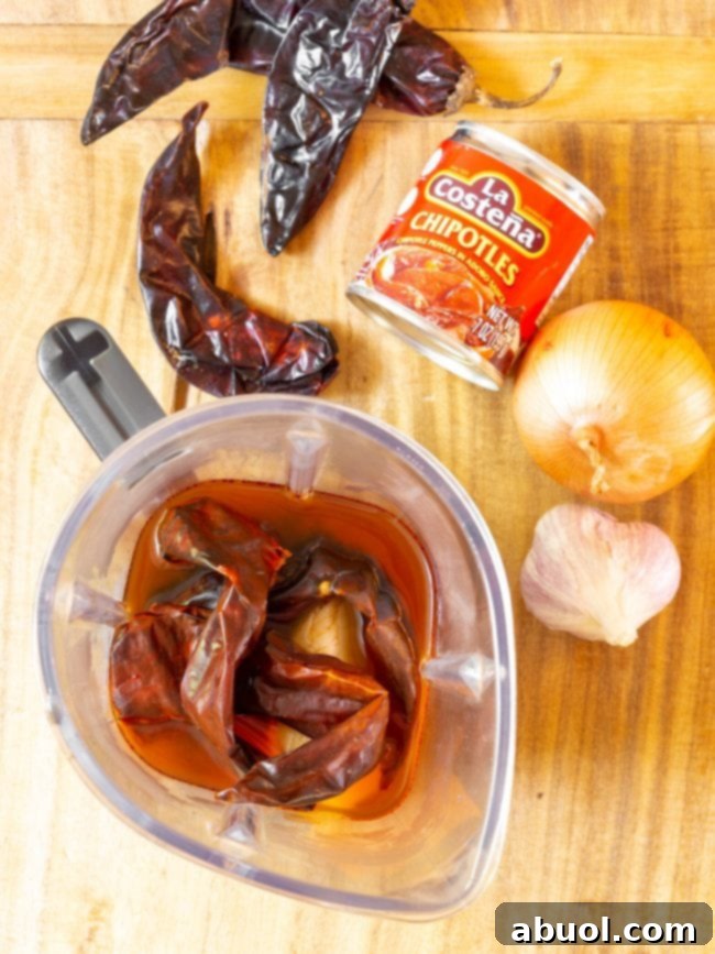 Essential ingredients for Crockpot Barbacoa: a can of chipotle peppers in adobo, dried New Mexico chiles, a whole sweet onion, a bulb of garlic, and peppers soaking in liquid in a blender jar, ready to be pureed.