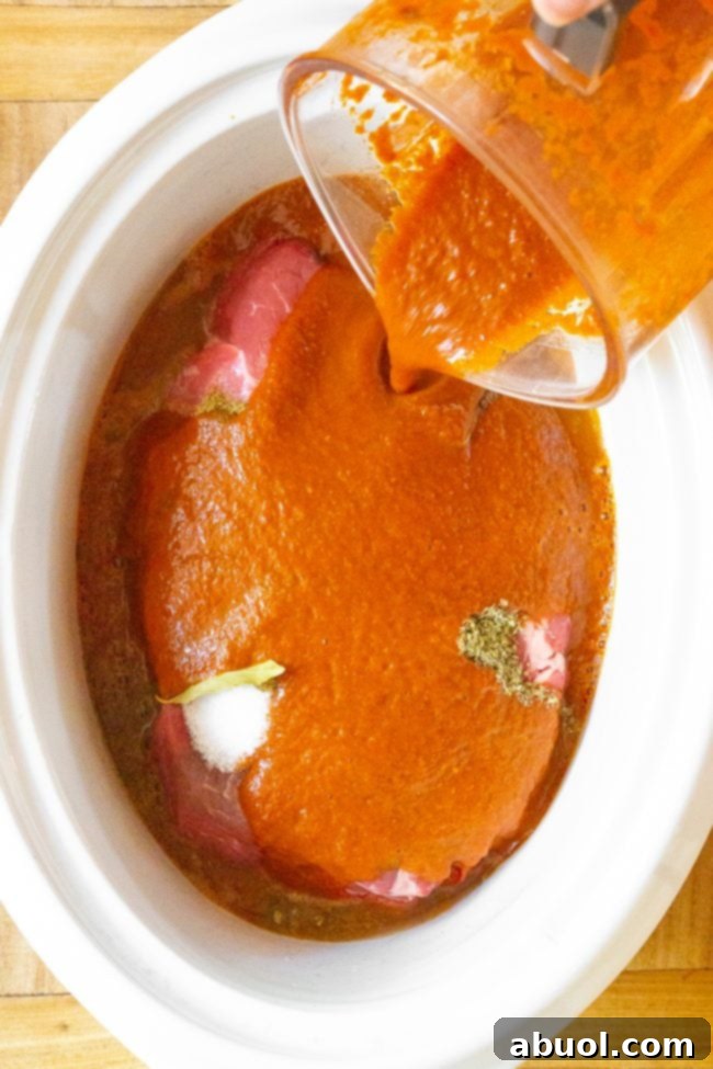Vibrant red spicy chile sauce being poured from a blender into a slow cooker over beef and barbacoa seasonings.