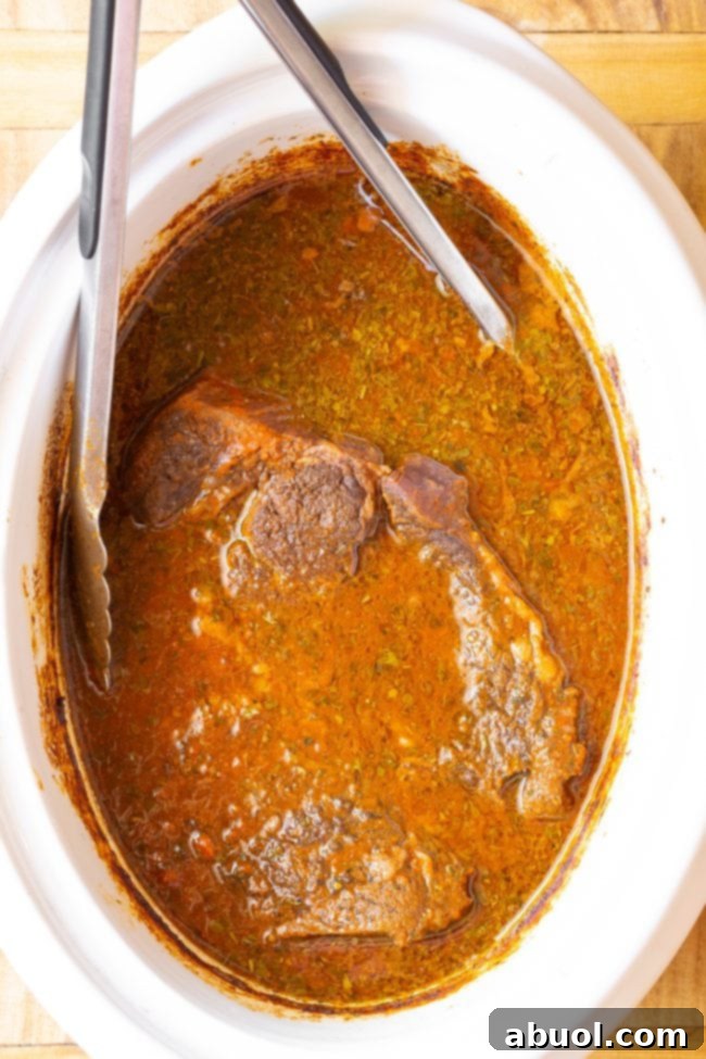 Metal tongs gently shredding tender, saucy beef directly into the vibrant red chile sauce within the slow cooker.