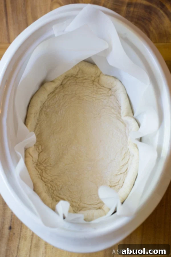 A top down view of raw pizza dough meticulously pressed into the bottom and up the sides of an oval slow cooker lined with parchment paper.