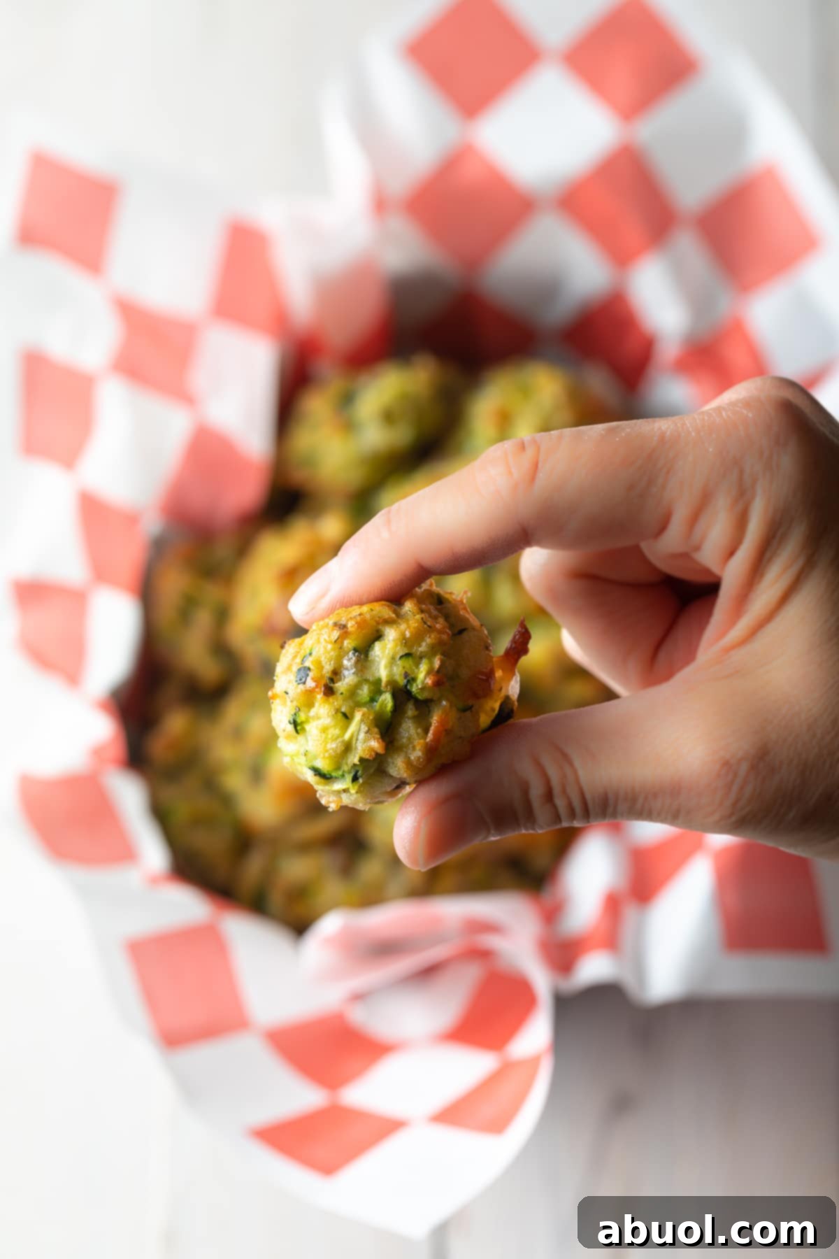 A hand holding a single perfectly golden and crispy baked zucchini tot towards the viewer, with more tots artfully blurred in the background, inviting a delicious bite.