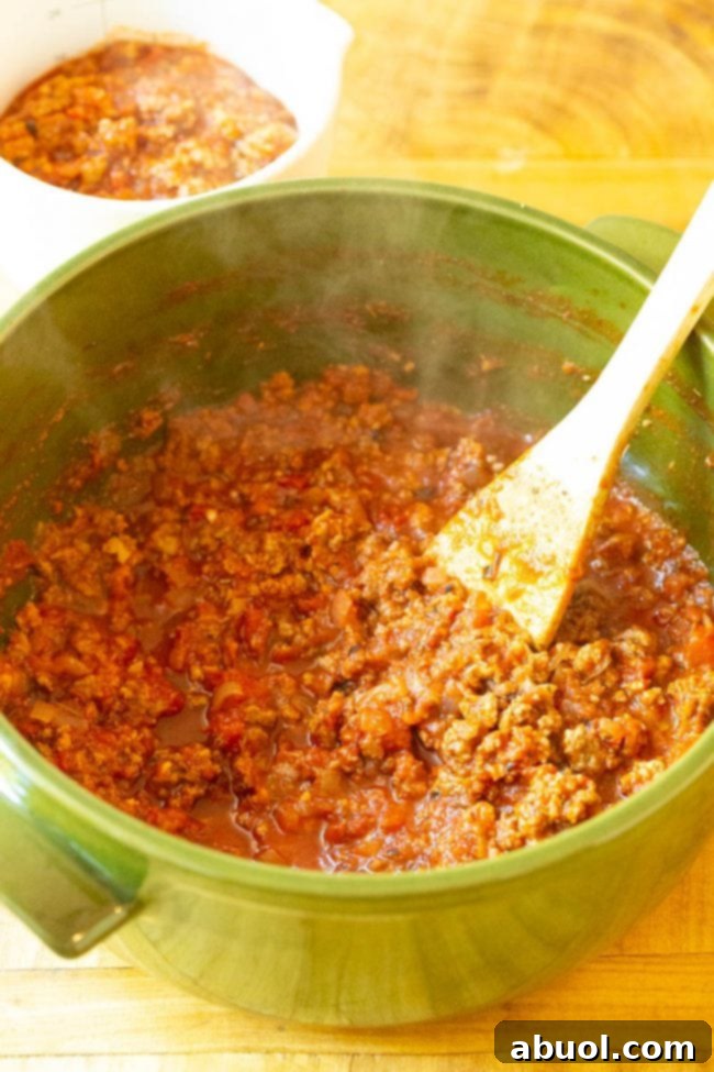 Close-up of raw ground beef and Italian sausage browning in a pan with chopped onions and minced garlic, the start of a rich meat sauce.
