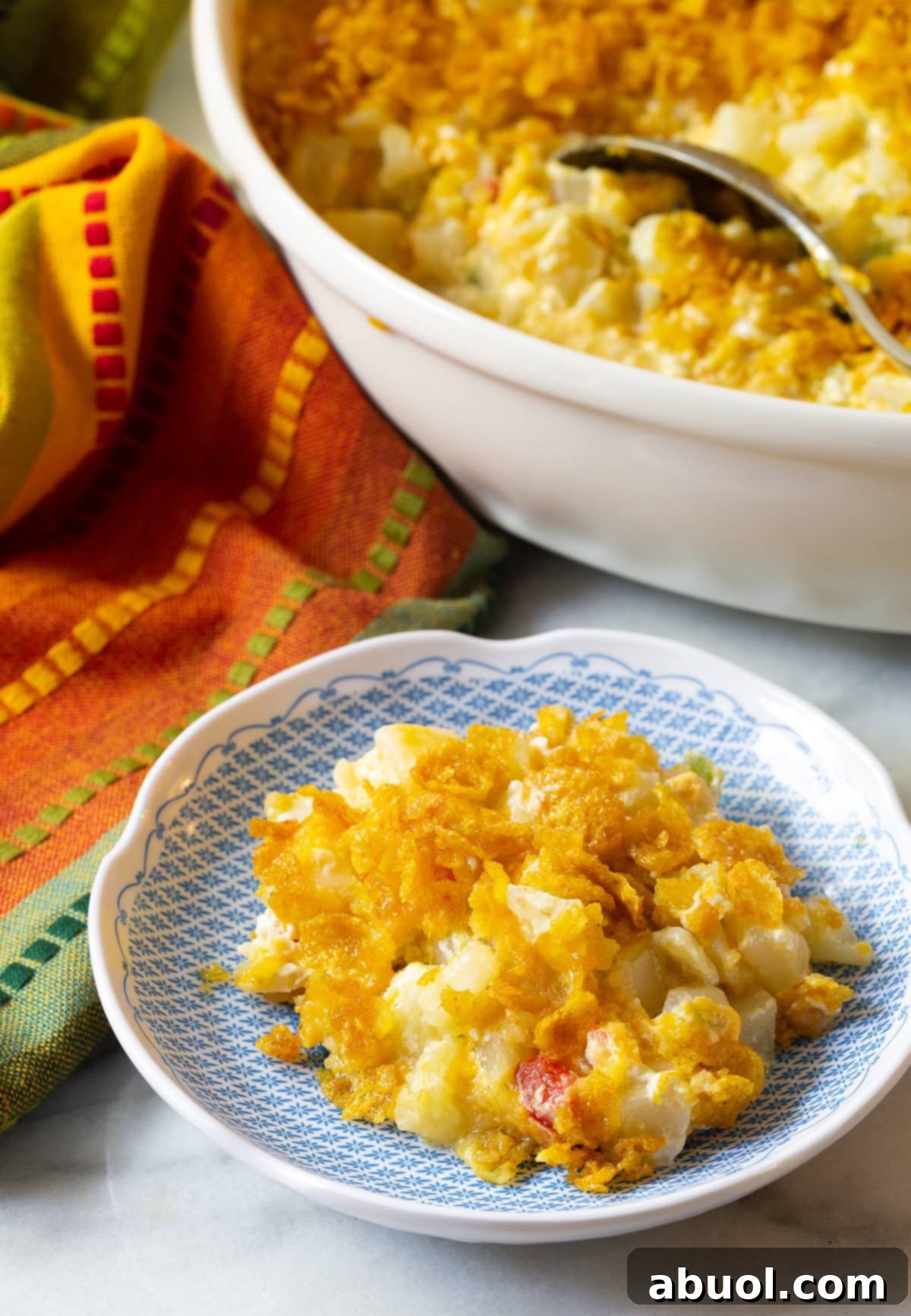 A serving of Company Potatoes with a crispy topping on a plate, with the full casserole dish in the background.