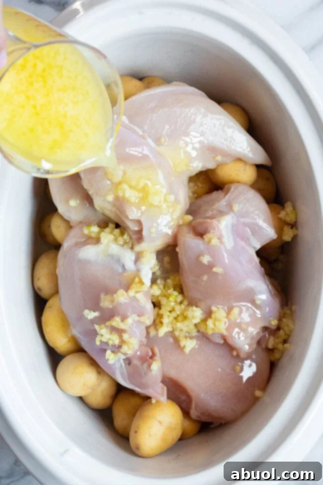 Raw chicken and potatoes in a slow cooker with a garlic butter mixture being poured over the top. 