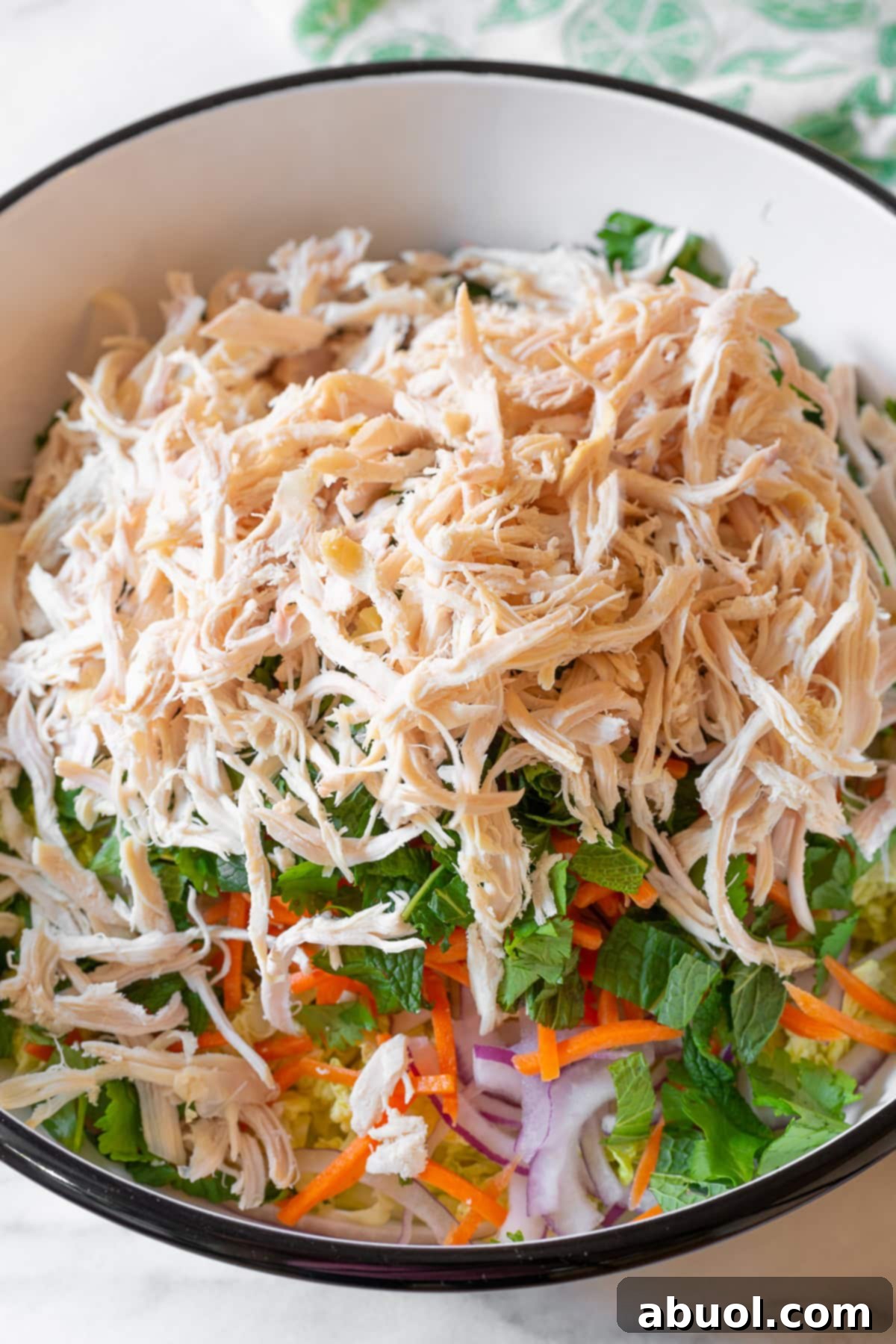 Shredded cooked chicken breast layered on top of the colorful mix of fresh vegetables in a large mixing bowl, awaiting the dressing.