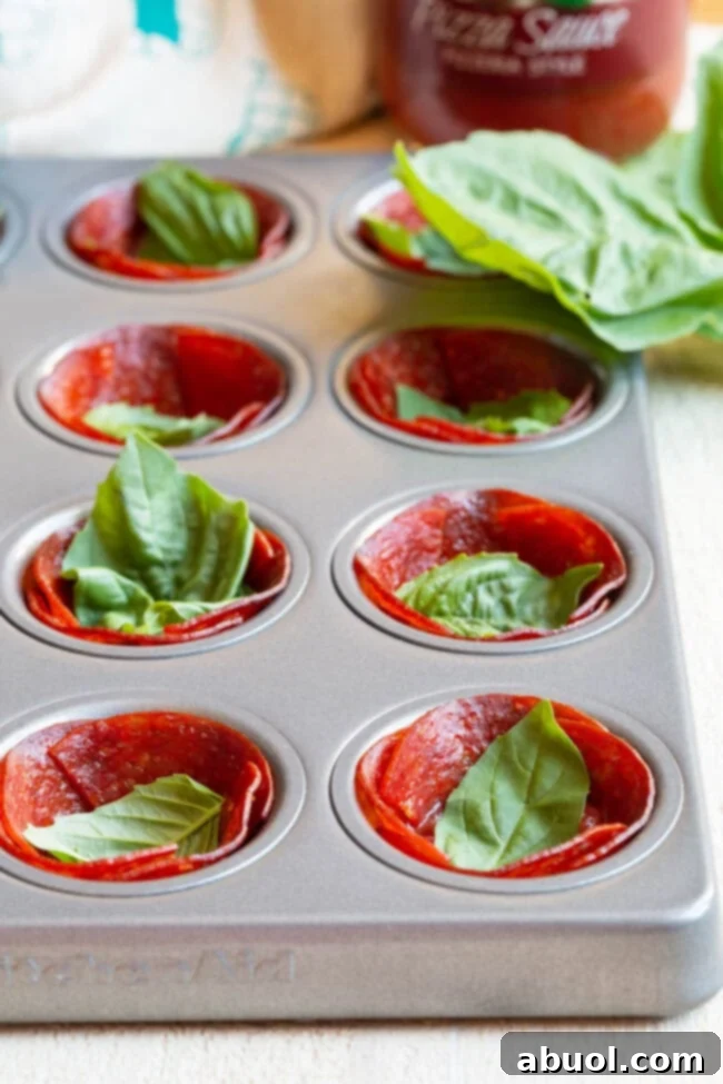 Basil Base for Pepperoni Cups Pepperoni cups in a muffin tin, each with a fresh basil leaf at the bottom.