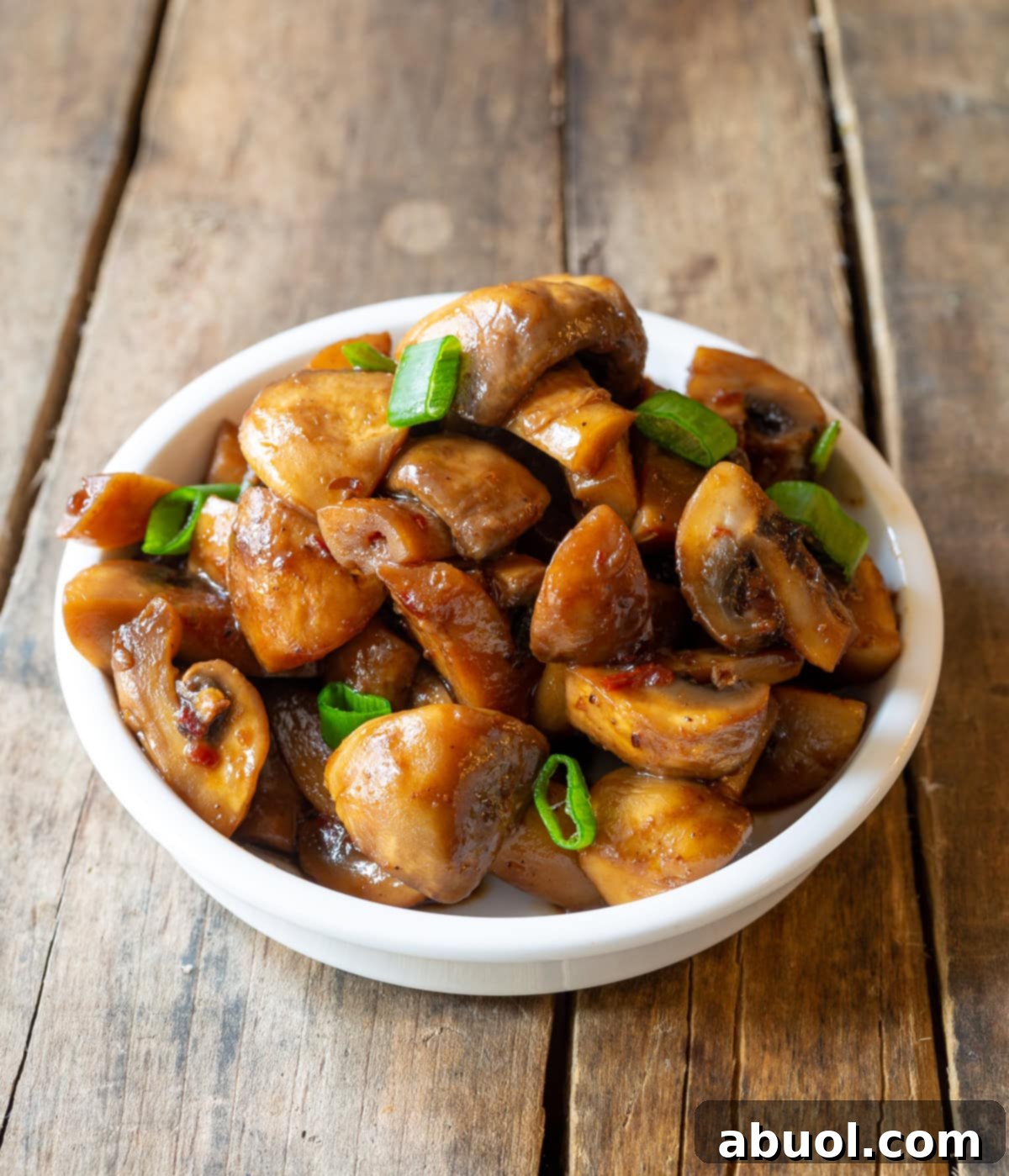 A crisp white bowl filled with glistening stir-fried Asian mushrooms, garnished with fresh green scallions, ready to be served. The rich brown tones of the mushrooms suggest deep flavor.