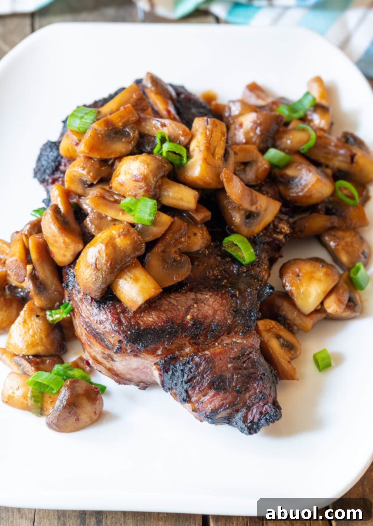 A perfectly cooked steak on a white plate, generously topped with glossy stir-fried Asian-style mushrooms and fresh scallions, creating an appealing main course.