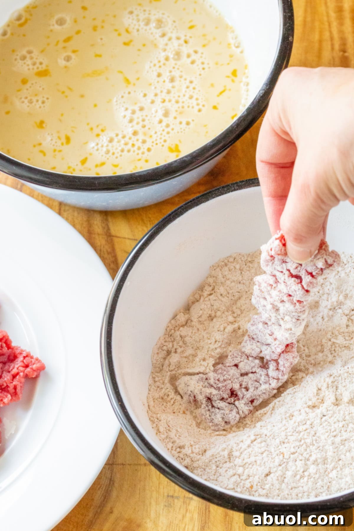 A pair of hands carefully dips a raw beef strip into a bowl of seasoned flour, preparing it for the frying process to create crispy steak fingers.