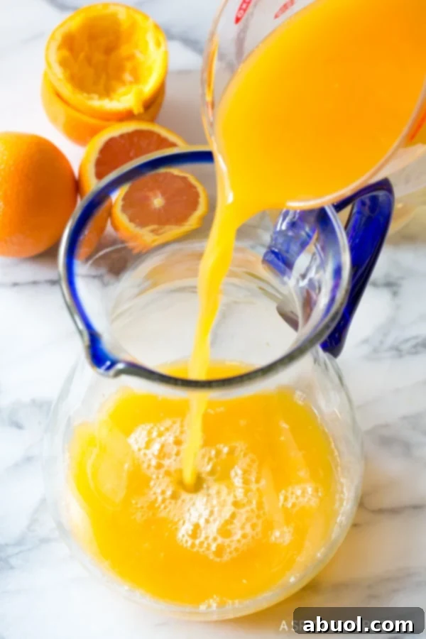 Freshly squeezed, golden orange juice being poured from a glass measuring cup into a large glass pitcher, signifying the start of orangeade preparation.