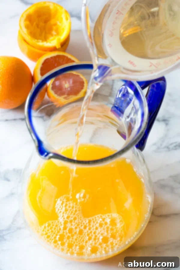 Clear simple syrup cascading from a measuring cup into a glass pitcher, mixing with the existing orange juice base for orangeade.