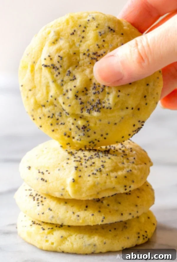 A hand holding a perfectly baked lemon poppy seed cookie, with more cookies stacked beneath, emphasizing its soft interior.