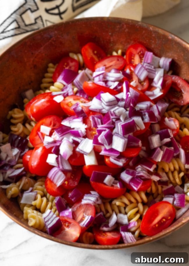 Variety of fresh ingredients for Italian pasta salad including cherry tomatoes, red onion, Italian parsley, mini mozzarella balls, and black olives laid out on a cutting board.