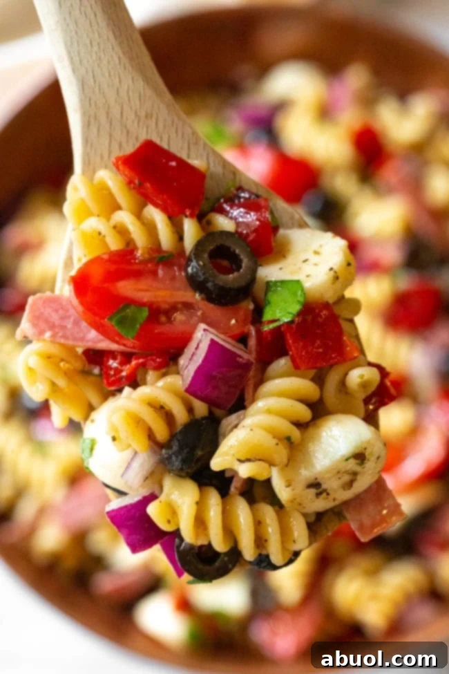 A wooden serving spoon holding a generous scoop of Italian pasta salad, highlighting the variety of pasta, vegetables, salami, and mozzarella in each bite.