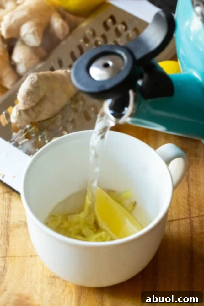Hot water being poured over grated ginger and lemon. A stream of hot water being poured from a kettle into a mug containing grated ginger and a lemon wedge.