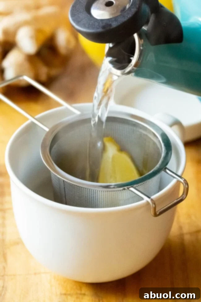 Pouring hot water over the lemon and ginger in a tea infuser in a mug. A close-up shot of hot water being poured into a mug with a tea infuser basket containing fresh ginger and lemon.