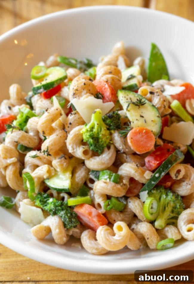 A white bowl filled with Skinny Pasta Primavera, featuring colorful chopped zucchini, carrots, broccoli, and red peppers, garnished with shaved parmesan cheese.