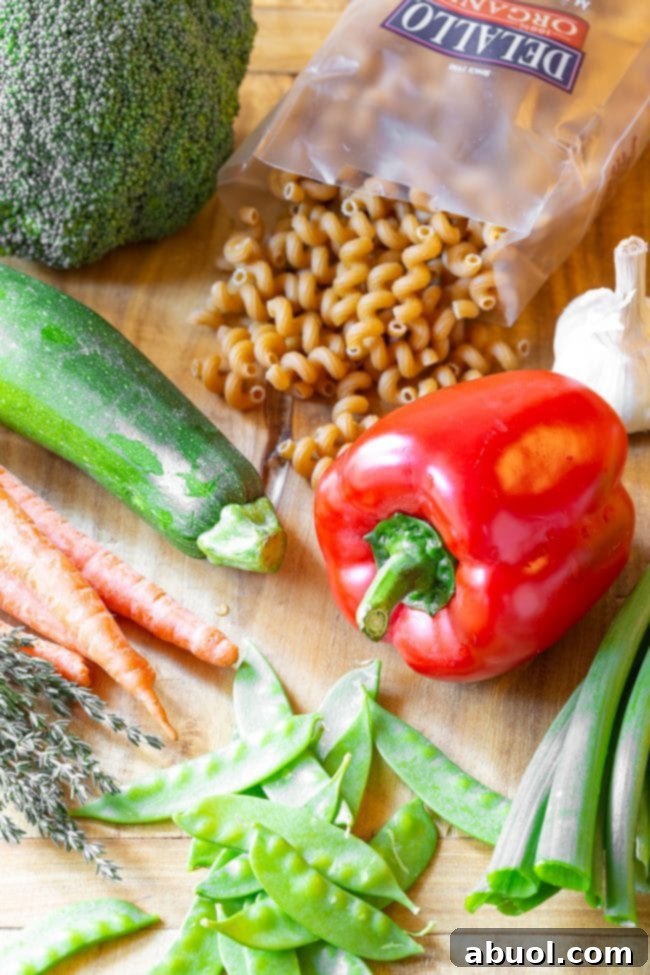 A vibrant display of fresh recipe ingredients: a whole red bell pepper, zucchini, carrots, snow peas, green onions, raw fusilli pasta spirals, and a bulb of garlic.