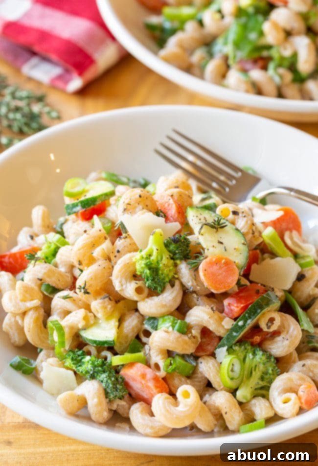 A white bowl of creamy noodles generously mixed with chopped zucchini, carrots, broccoli, red peppers, and topped with delicate shaved parmesan cheese, showcasing its rich texture.