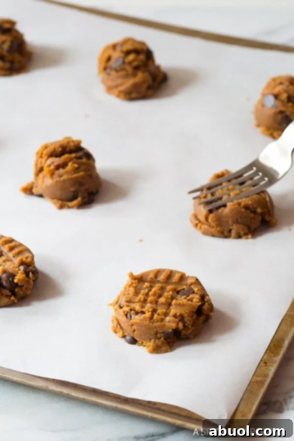 A fork gently pressing down on a ball of cookie dough on a baking sheet, creating the classic crosshatch pattern before baking.