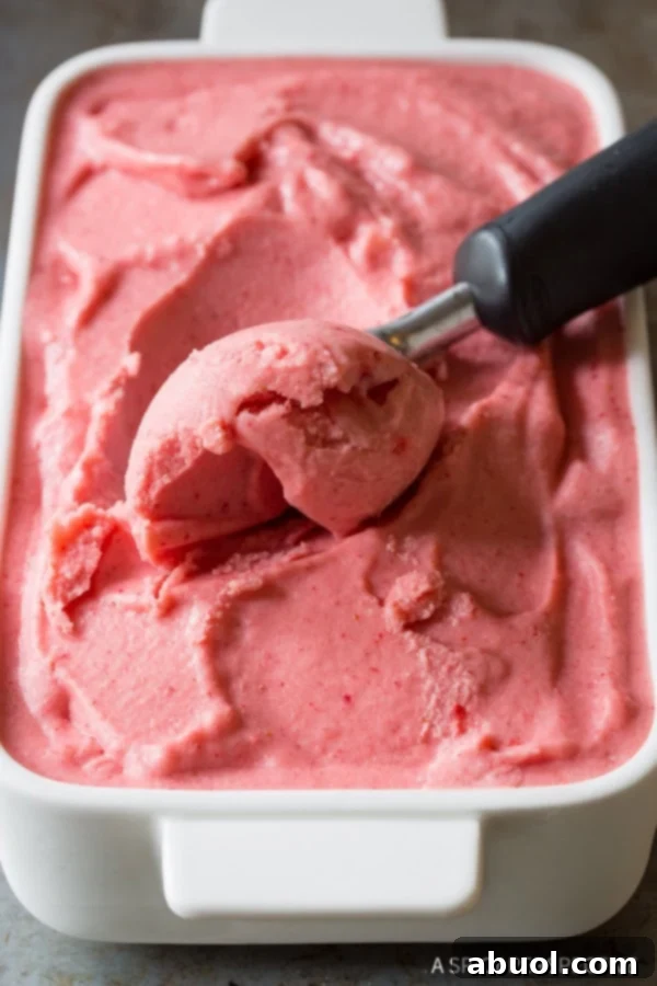 An ice cream scoop delicately carving a perfect sphere of strawberry pineapple sherbet from a pristine white serving dish.