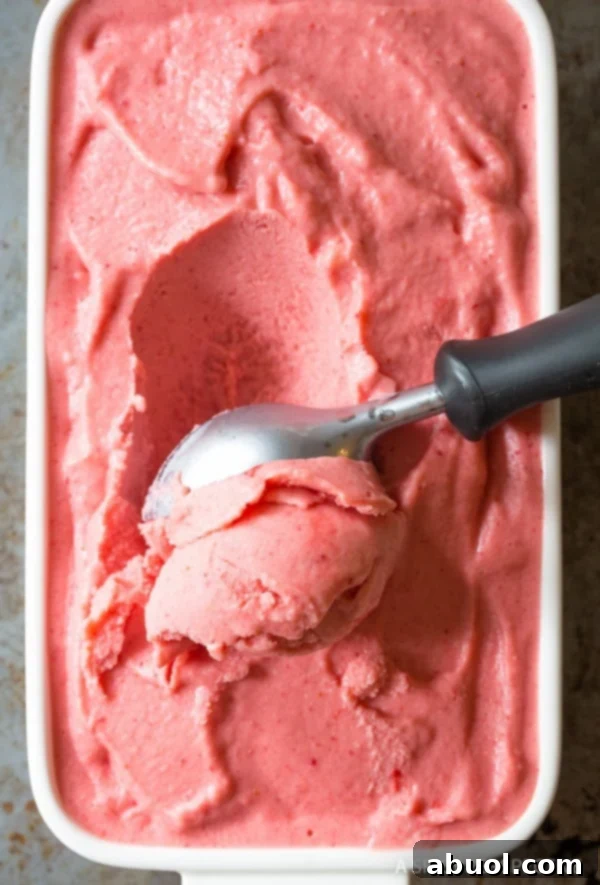A top-down view of an ice cream scoop effortlessly scooping creamy strawberry sherbet from a white serving dish, revealing its delightful texture.