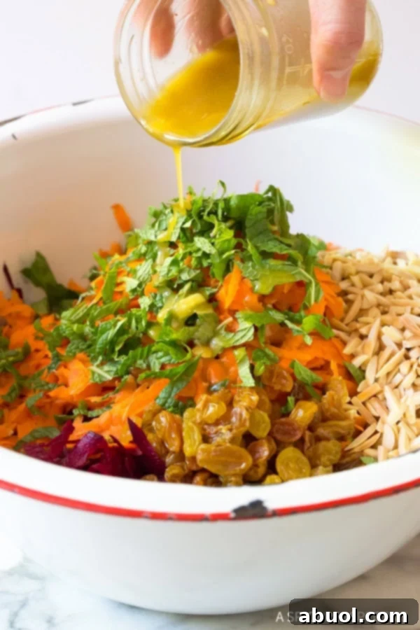 A hand pouring dressing over shredded carrots, beets, mint, sultanas and almonds in a bowl, mid-toss.