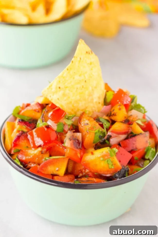 A vibrant closeup of a bowl filled with chunky grilled peach salsa, adorned with a single nacho chip ready for dipping.