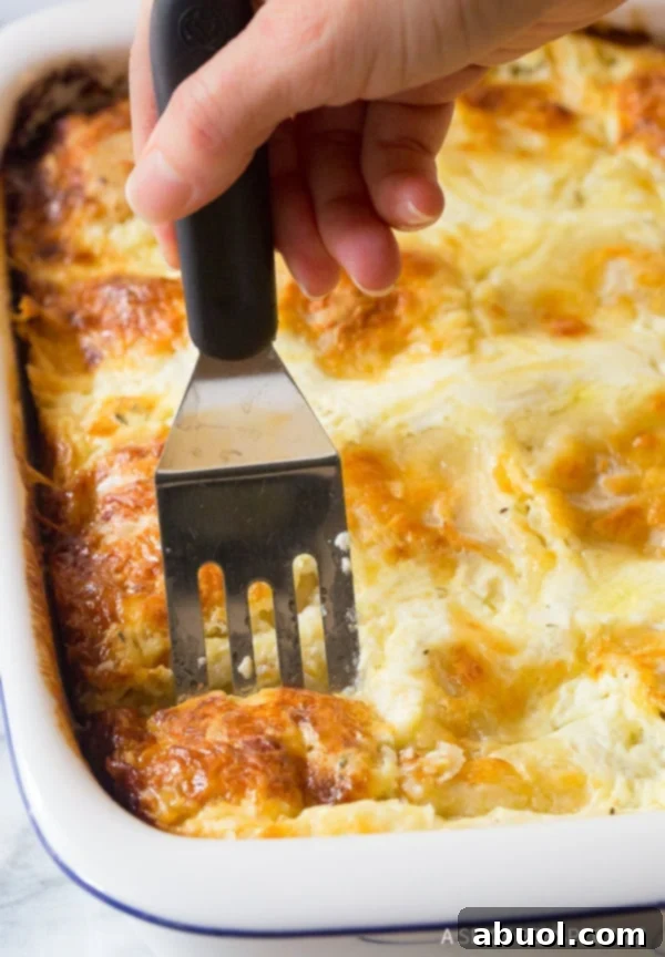 A hand gently lifts a steaming, golden-brown slice of Croatian Cheese Strukli from a large baking dish using a spatula, showcasing its layers of tender dough and creamy cheese filling.