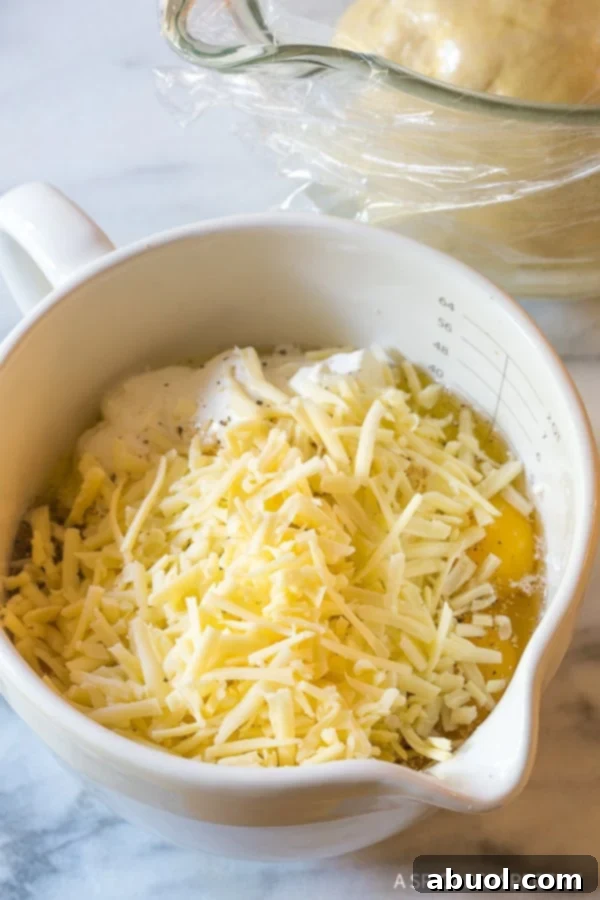 Finely shredded white cheddar cheese being added to the creamy cottage cheese, sour cream, and egg mixture in a glass mixing bowl, ready for the Croatian Cheese Strukli filling.