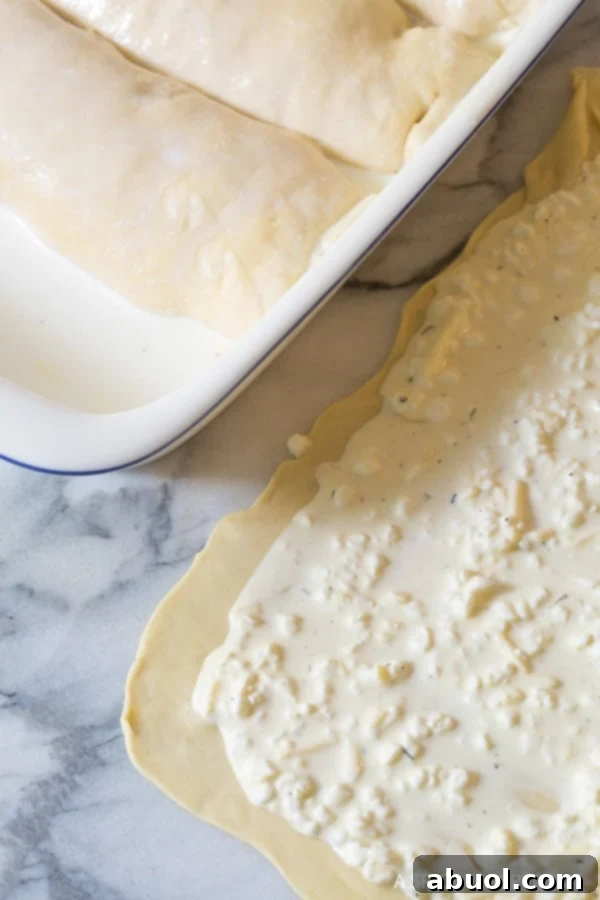 A thin sheet of homemade dough, generously covered with the creamy cheese mixture, is skillfully rolled into a log, a crucial step in preparing Croatian Cheese Strukli.