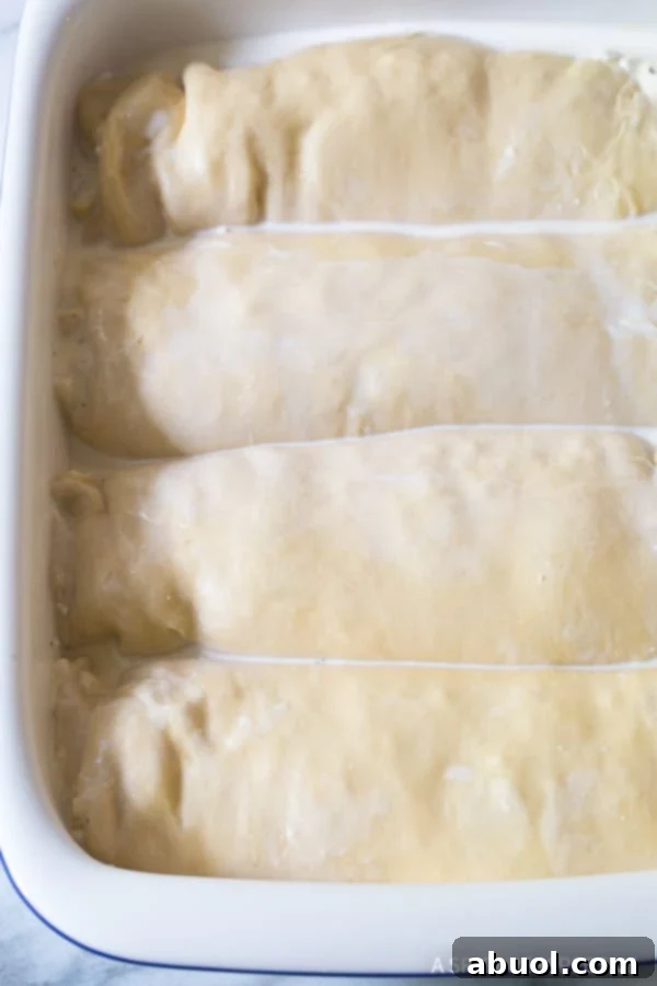 Four neatly rolled logs of Croatian Cheese Strukli dough, filled with a creamy cheese mixture, are arranged side-by-side in a large glass baking dish, ready for the next step of preparation.