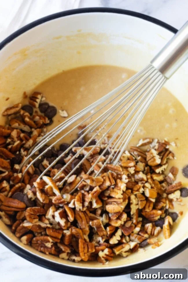 Hands whisking chopped pecans and dark chocolate chips into a rich, eggy pie batter in a large mixing bowl.