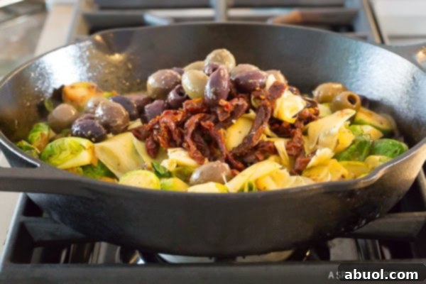 Adding sun-dried tomatoes, greek olives, and artichoke hearts to the cast iron skillet with brussel sprouts