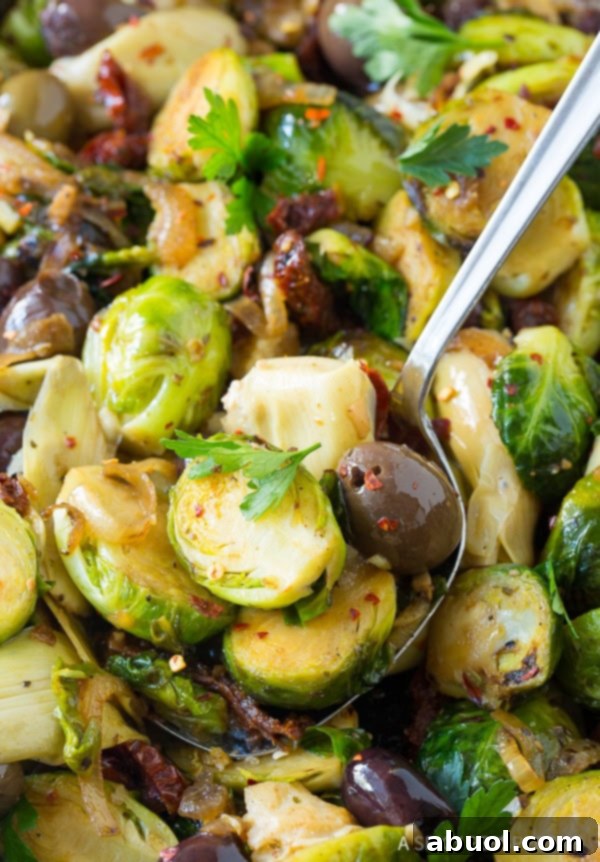 Metal spoon scooping up a servings of baised brussels sprouts with sun-dried tomatoes, greek olives, artichoke hearts and fresh chopped parsley.